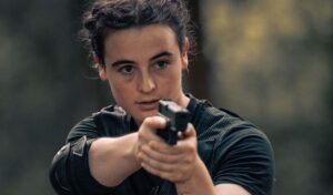 A woman with dark curly hair pulled back away from her face holds a shot gun out in front of her.