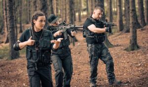 3 people walk through the woods in dark camo clothing. 2 men are carrying machine guns while a woman demonstrates the correct way to hold them.