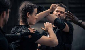 A woman and man in black T shirts and protective joint pads are mid fight - their forearms hit each other in the air