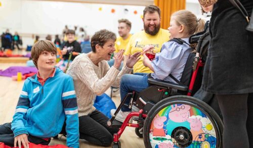 A young girl sits in a wheelchair laughing and playing with two workshop practioners On the left hand side a young boy wearing a blue jumper sits on the floor looking up at someone off camera