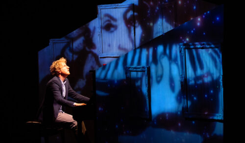An image of a man playing the piano There is a set behind him featuring a womans face on a wooden panel under blue lighting