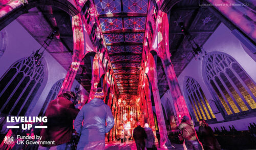 A picture of Southwell minster full of people The buildings interior is lit up in pink and purple lights In the bottom right are three logos which reads Levelling Up and Funded by UK Government