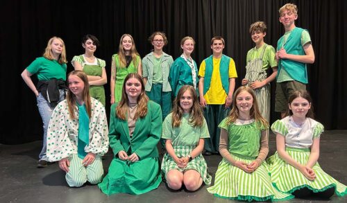 A group of young people dressed in bright green costumes A row of people stand on a stage in front of a black curtain In front of them is a row of people kneeling on the floor
