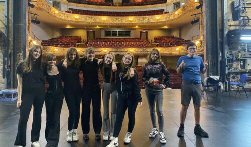 8 members of the Courtyard Youth Theatre stand on a stage with their backs to an auditorium full of red seats