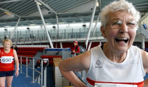 An elderly woman stands with her hands on her hips on an indoor athletics track