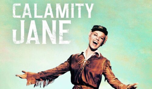 A woman in a brown jacket sings with her arms outstretched The text reads Calamity Jane