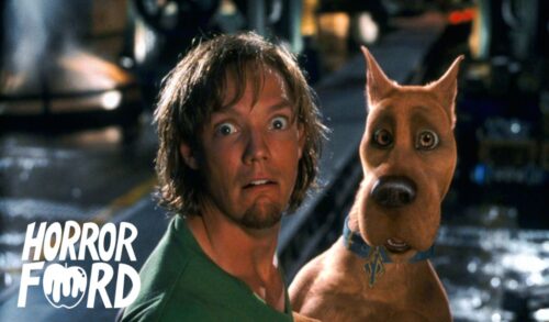 A man with long hair and a green shirt stands next to a CGI dog In the bottom left is the Horrorford logo