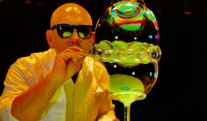 A bald man wearing sunglasses and a white shirt is blowing a series of large bubbles