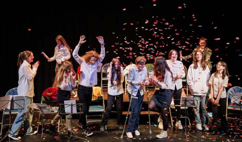 A group of young performers stand on stage celebrating Behind them is multicoloured confetti