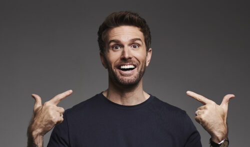 Joel Dommett stands smiling at the camera pointing at his face