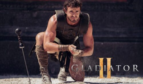 A man dressed as a Gladiator kneels on one leg and rubs dirt into his hands