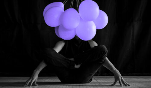 A person sits cross legged on the floor A bunch of purple balloons covers their face