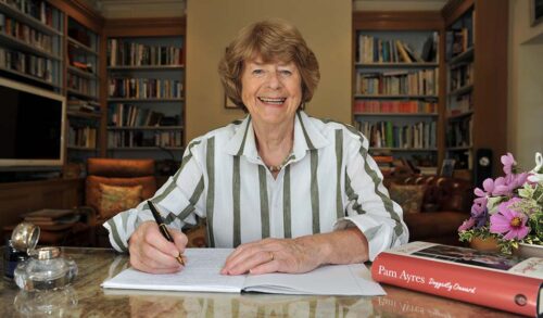 Pam Ayres sits at a table with an open book writing