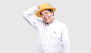 Mark Steel - the comedian Mark Steel is laughing, wearing a yellow hat and white shirt. His right arms is raised to his hat.