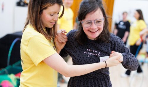 A woman with short dark hair wearing a bright yellow tshirt is holding the hands of a young girl who has special educational needs