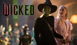 A green witch in a black cloak and hat stands next to another witch in a pink dress. In the top left corner is the 'Wicked' logo.