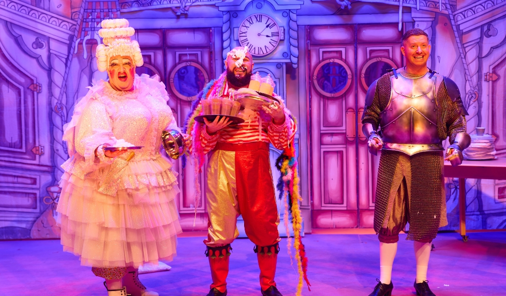 A pantomime Dame holds out a plate. Next to her a character with cream on his face holds two full trays. A character in a a suit of armour stands next to them.