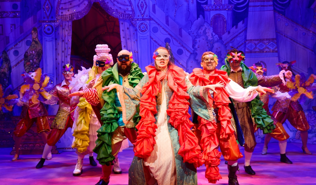 The pantomime cast dance on stage dressed in coloured scarves and masks.