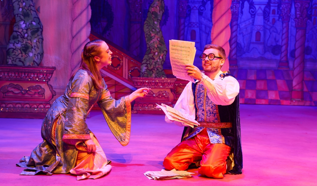 An actress dressed as Sleeping Beauty kneels on the floor pointing to a male character who holds up sheet music.