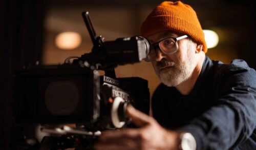 A bearded man wearing glasses and a yellow hat is operating a film camera