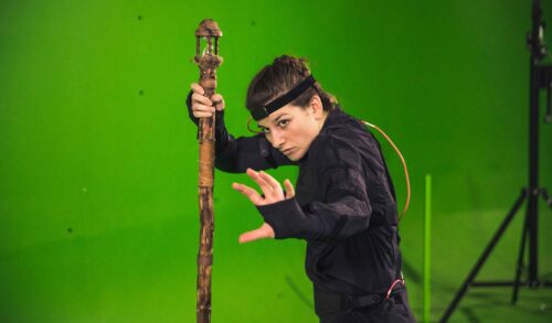 A woman wearing all black and motion capture equipment stands holding a wooden staff in one hand The other hand is reaching towards the camera