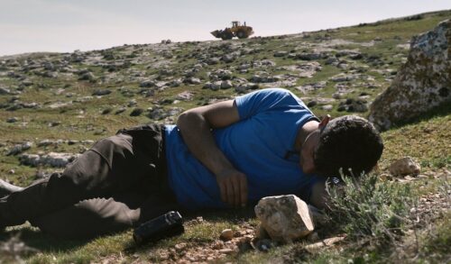 A man wearing a blue tshirt and black trousers lies on the ground of a mountainous terrain