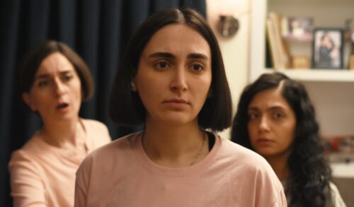 A woman with a bob haircut wearing a pink tshirt stands in front of two other woman who are looking concerned