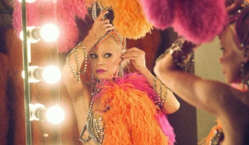 A close up of a woman covered in brightly coloured feather pinning a headdress to her head whilst looking in the mirror