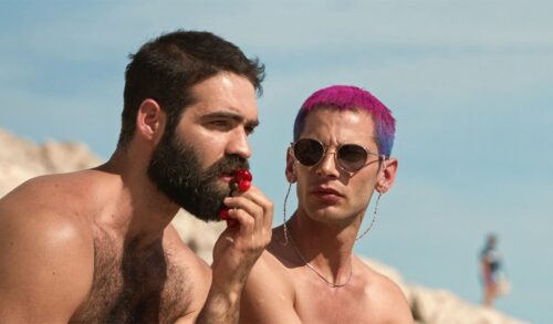 The Summer With Carmen  two men at the beach without tops on One has a dark beard and is eating a strawberry the other has pink hair and is wearing sunglasses