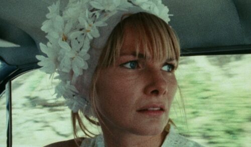 Wanda  a blonde woman in a 1960s hat with flowers on looks worried