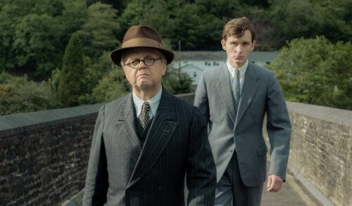 An older man in a suit and hat walks ahead of a younger man in a suit