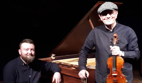 Tom McConville And Michael Biggins stand infront of a piano